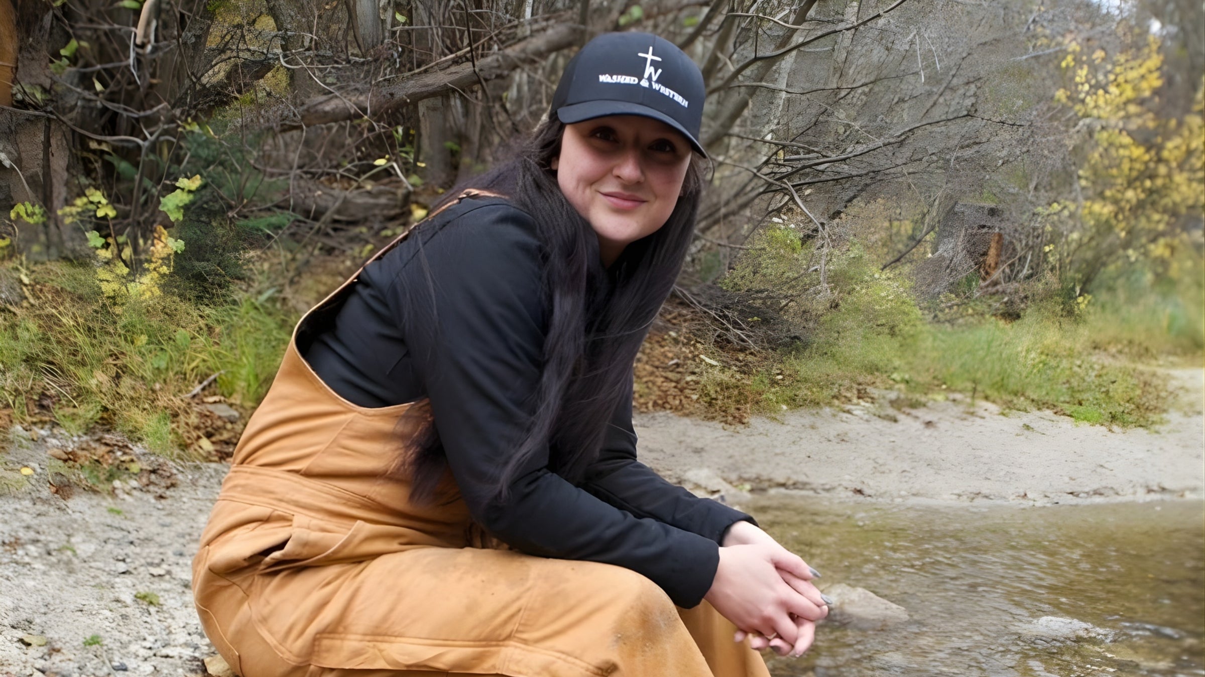 Woman sitting in overalls with a black Washed & Western cap on