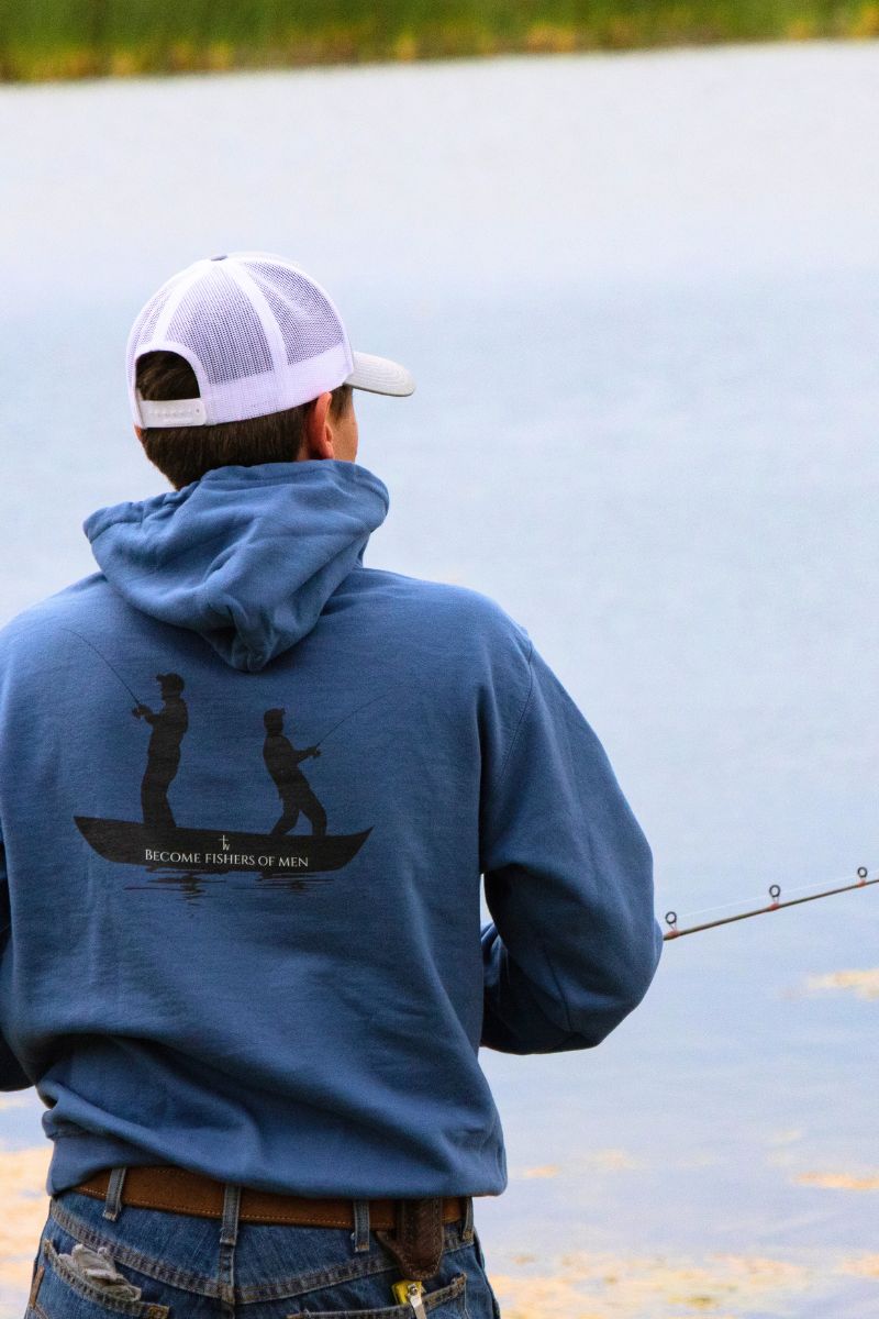 Person fishing by a lake wearing a blue hoodie with a graphic design.